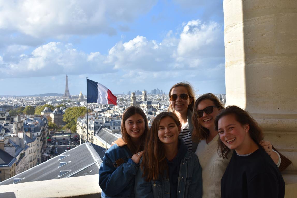 a group of students pose for a photo in front of a French flag and Paris backdrop, including the Eiffel Tower