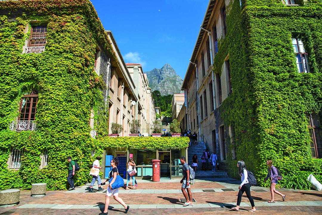 Students walk past buildings covered in lush vegetation on a sunny day at the University of Cape Town.