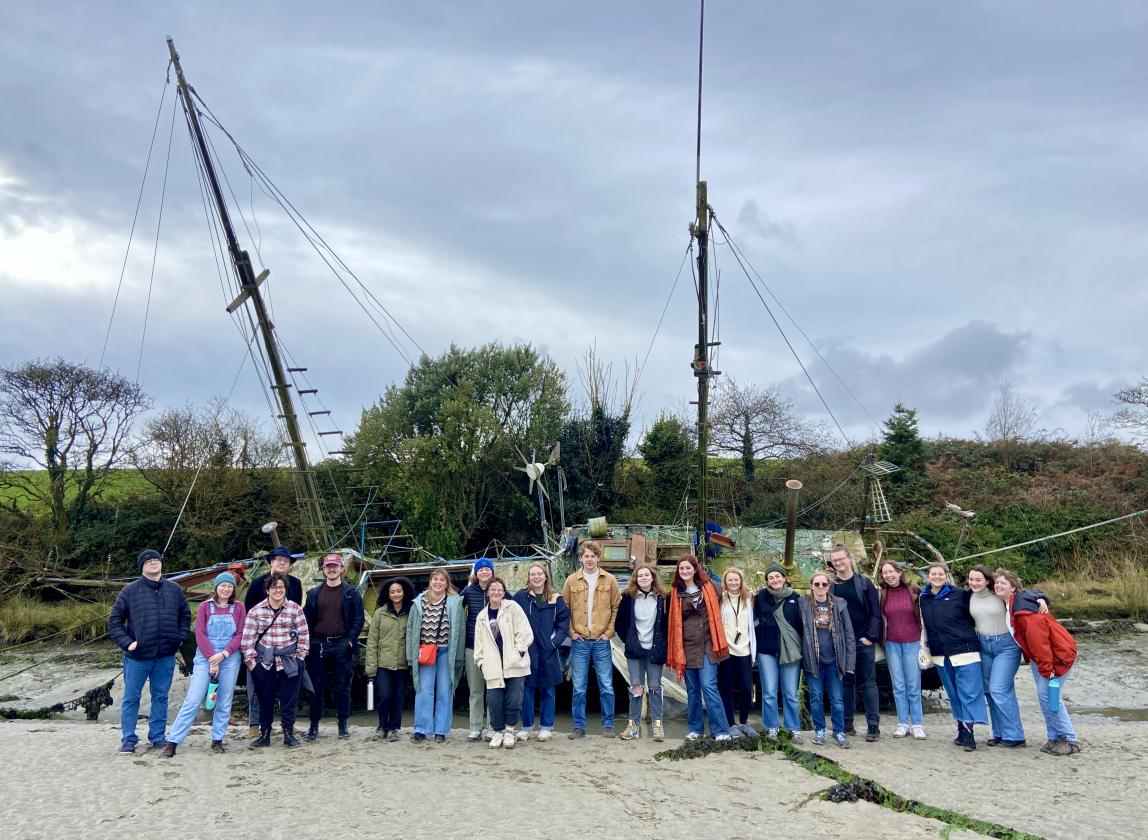 large group of students standing on a sand beach in front of a massive boat with grass, trees, and fog in the background