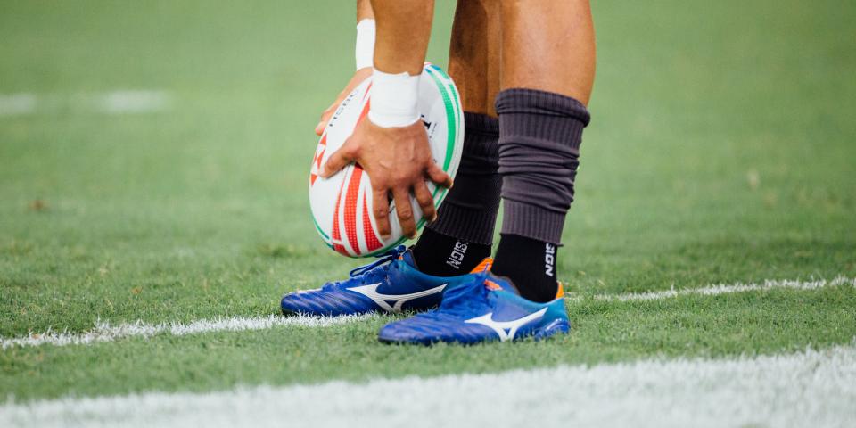 A man in rugby gear holds a rugby ball by his feet